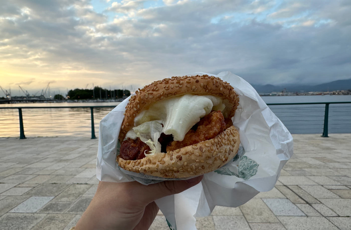 Hand håller i en burgare med ljust bröd, isbergssallad och friterad kyckling.