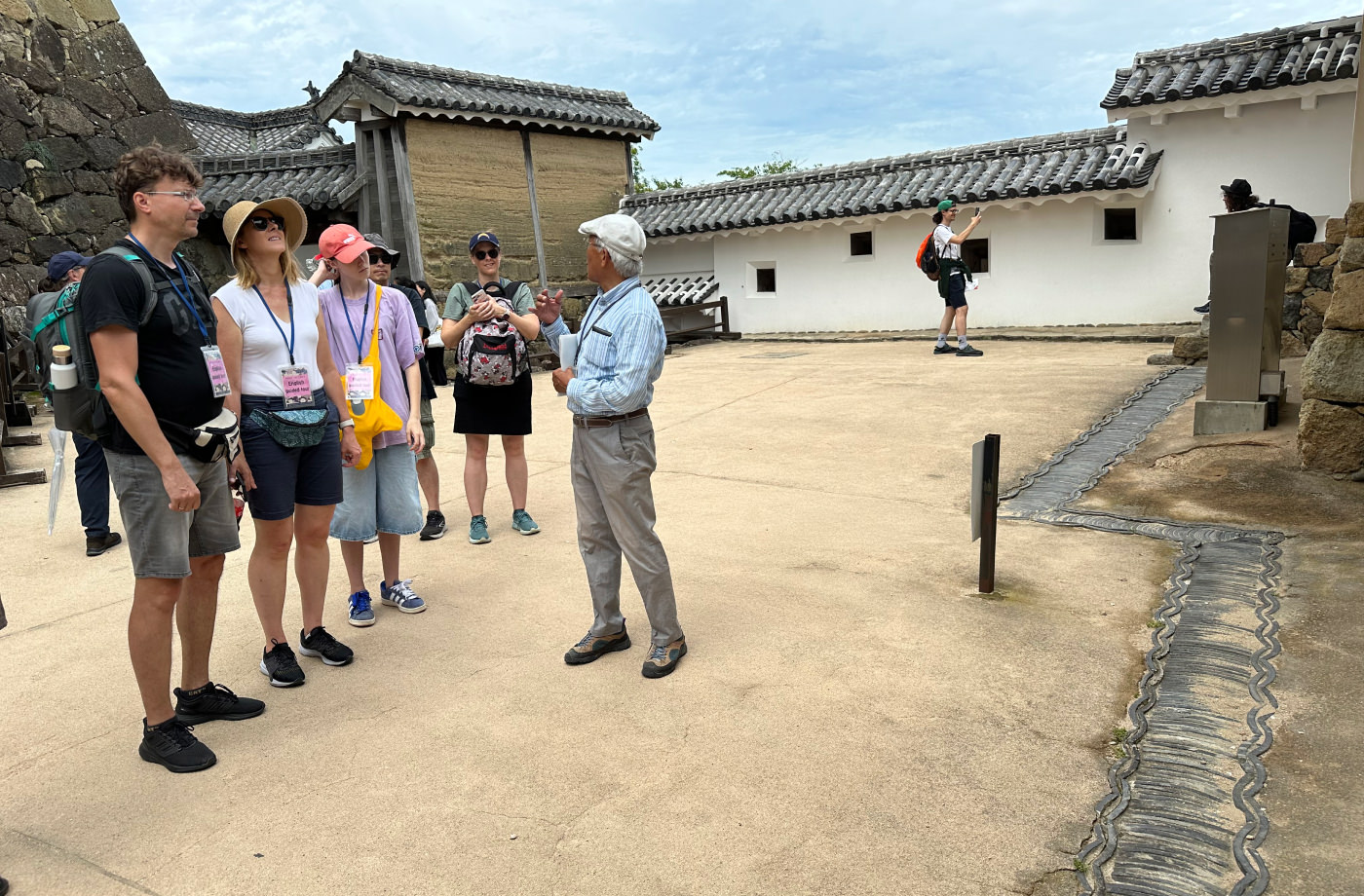 En grupp med människor står utomhus inne på en liten slottsgård och lyssnar på en guide. Till höger på marken syns ett gammalt avrinningssystem för regnvatten.