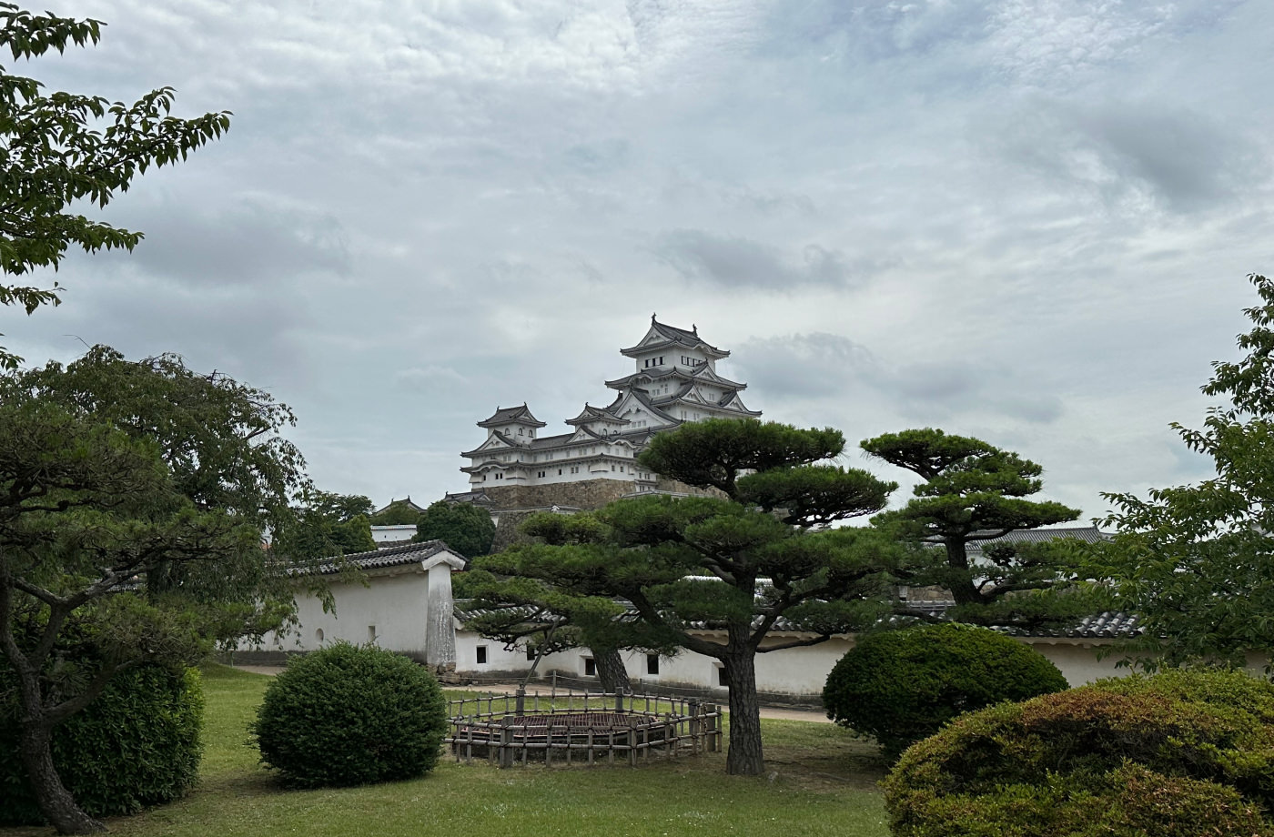 I fjärran syns ett sex våningar högt och vitt japanskt slott med gråa tak på flera torn. I förgrunden har vi en välansad trädgård.