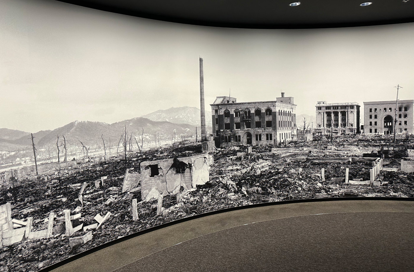 Stort svartvitt panoramafoto täcker en väg som böjer sig till höger. Fotot visar en sönderbombad stad med få byggnader som står kvar.