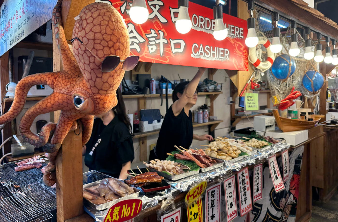 Grillspätt med fisk och skaldjur på en bardisk vid ett matställe. Till vänster hänger ett kramdjur av en bläckfisk. Kramdjuret har solglasögon på sig.