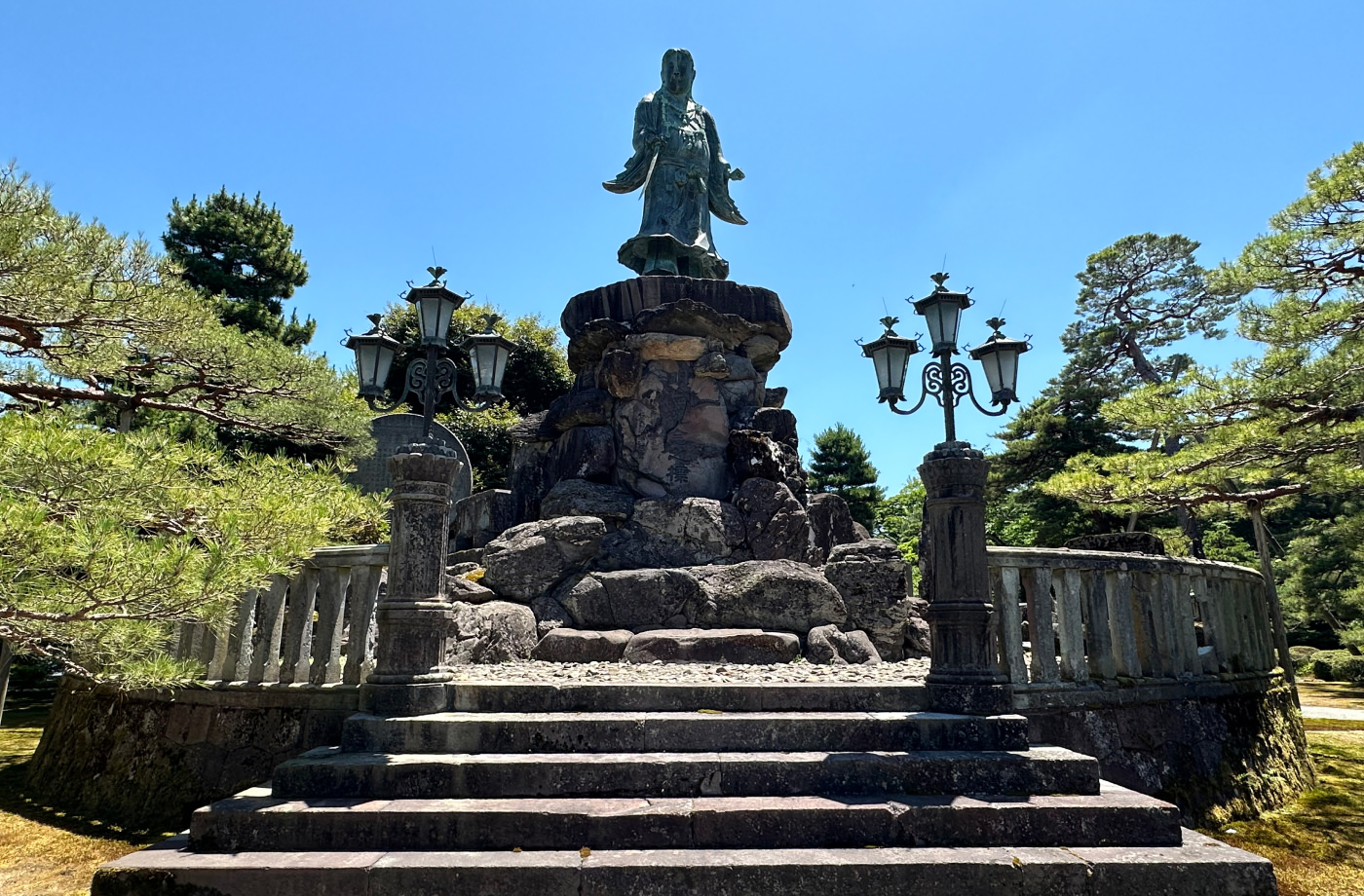 Staty på en man står på en hög stenpedestal på en rund platå med stenräcken och lyktor omgivna av buskar och träd.