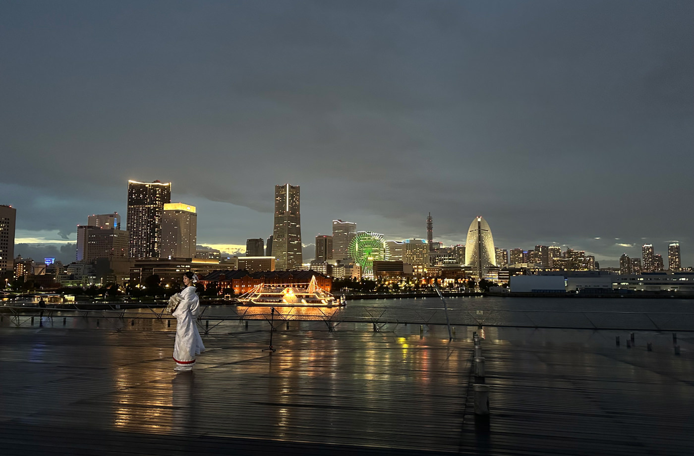 Ung japansk kvinna i vit traditionell bröllopsdräkt på ett blött trädäck. I fjärran en stor, vacker och upplyst skyline framför en hamn.