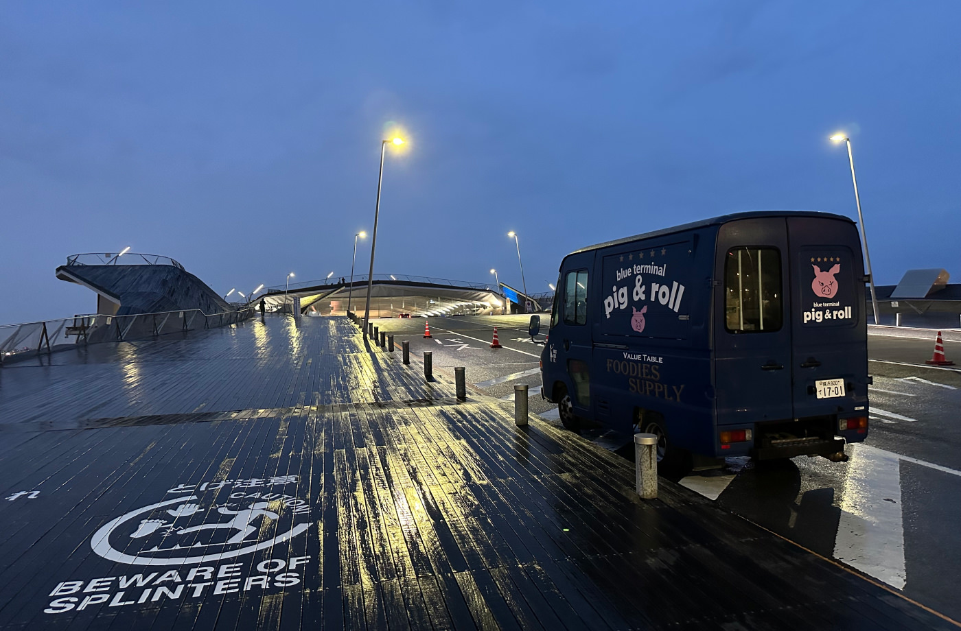 Backe upp till en stor färjeterminal. Till vänster är en blöt trätrottoar med texten "Bewsare of splinters". Till höger stör en mörkblå skåpbil med texten "Blue Terminal. Pig & Roll".