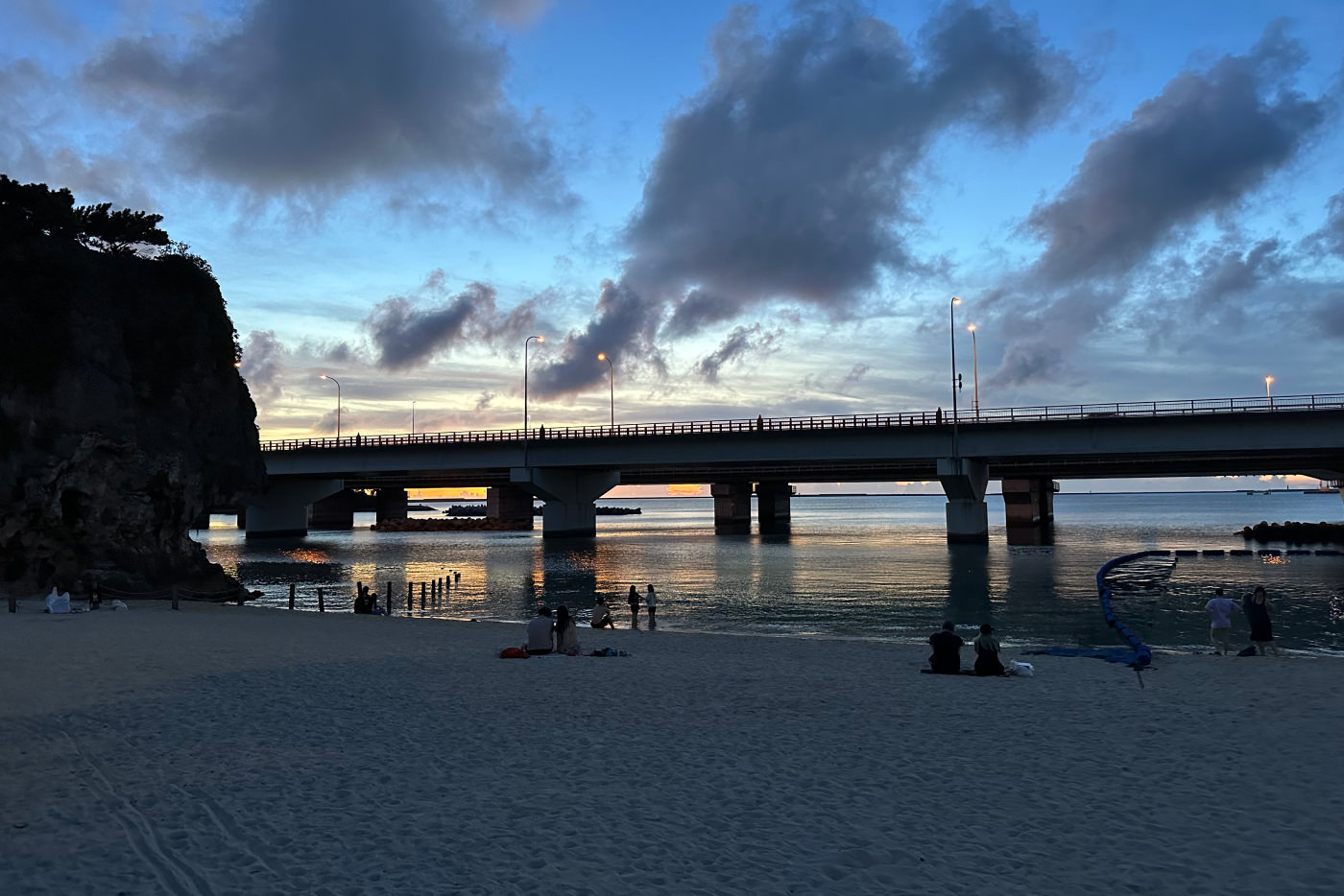 Liten sandstrand framför en cementbro med tända lyktstolpar på. Solen går ned och reflekteras i vattnet. Drygt tio personer är på plats.