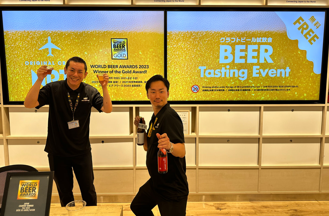 Två japanska män i svarta kläder poserar med ölglas och ölflaskor framför två gula TV-skärmar. På en står det "Beer Tasting Event".