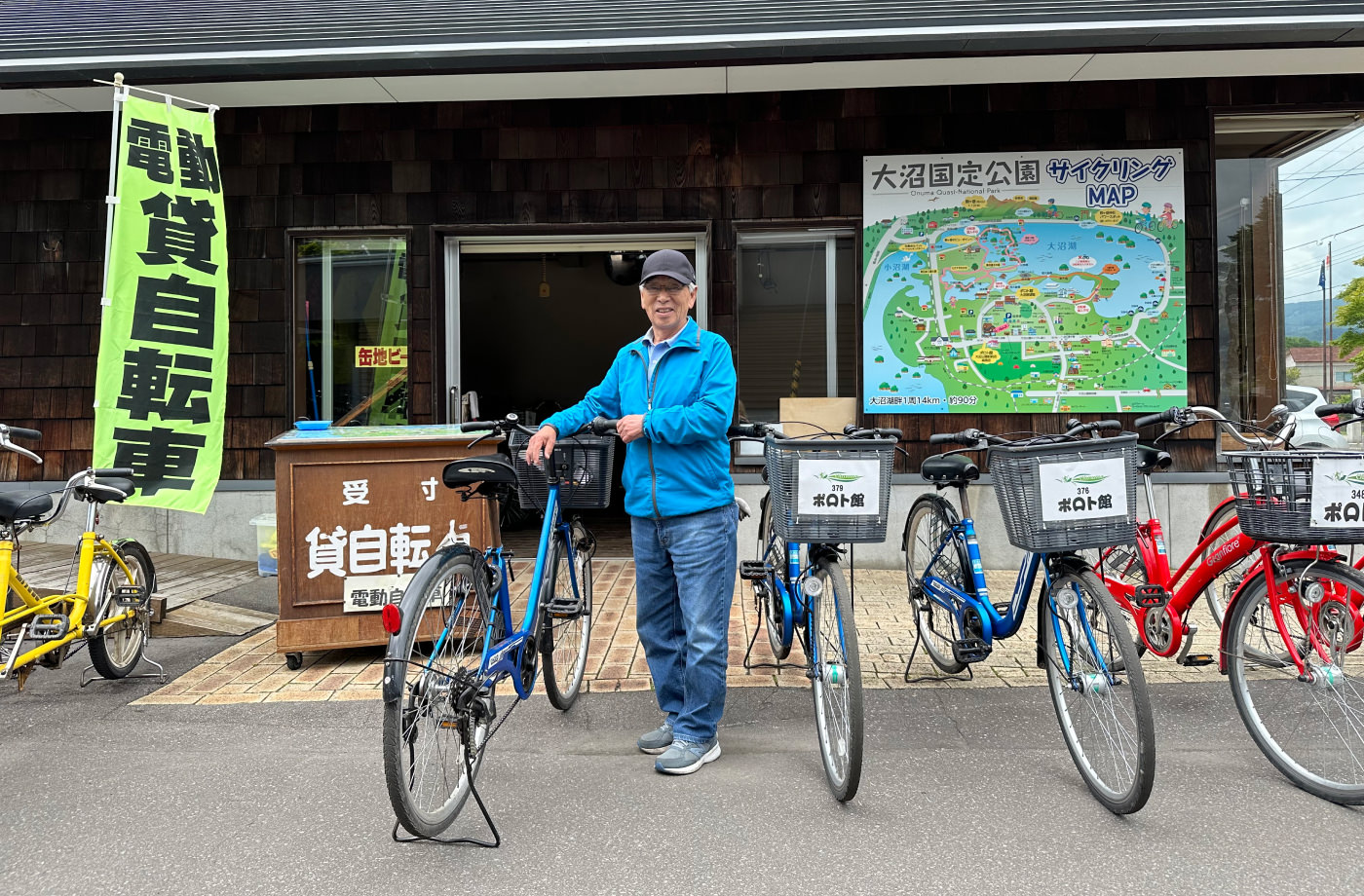 Äldre japansk man i blåa kläder och keps står vid några uppradade cyklar.