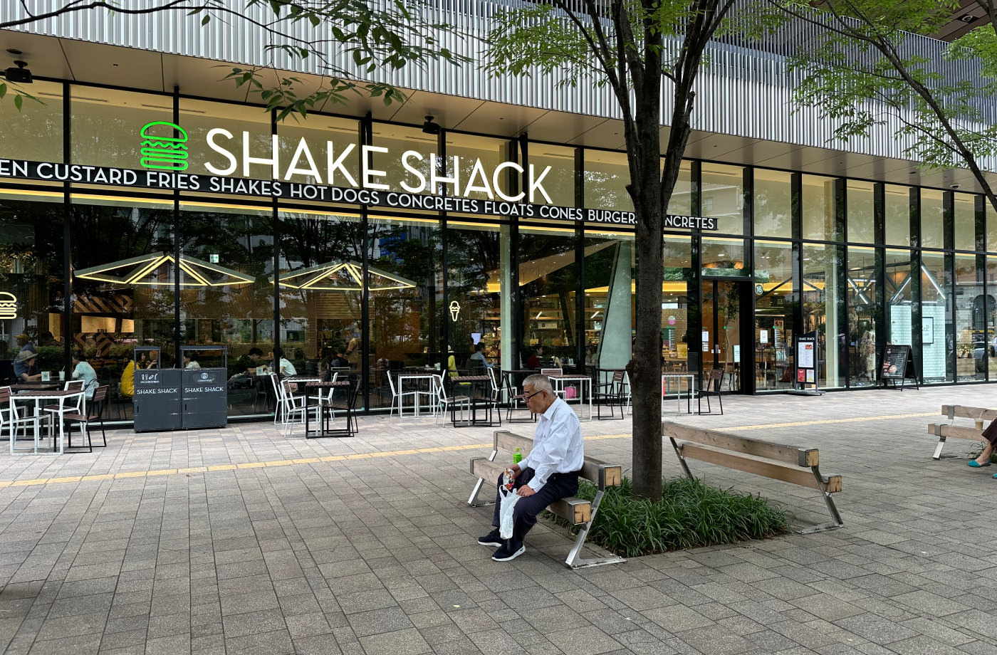 En äldre man sitter i en liten park framför en stor glasfasad med titeln "Shake Shack" efter en grön neonskylt av en hamburgare.