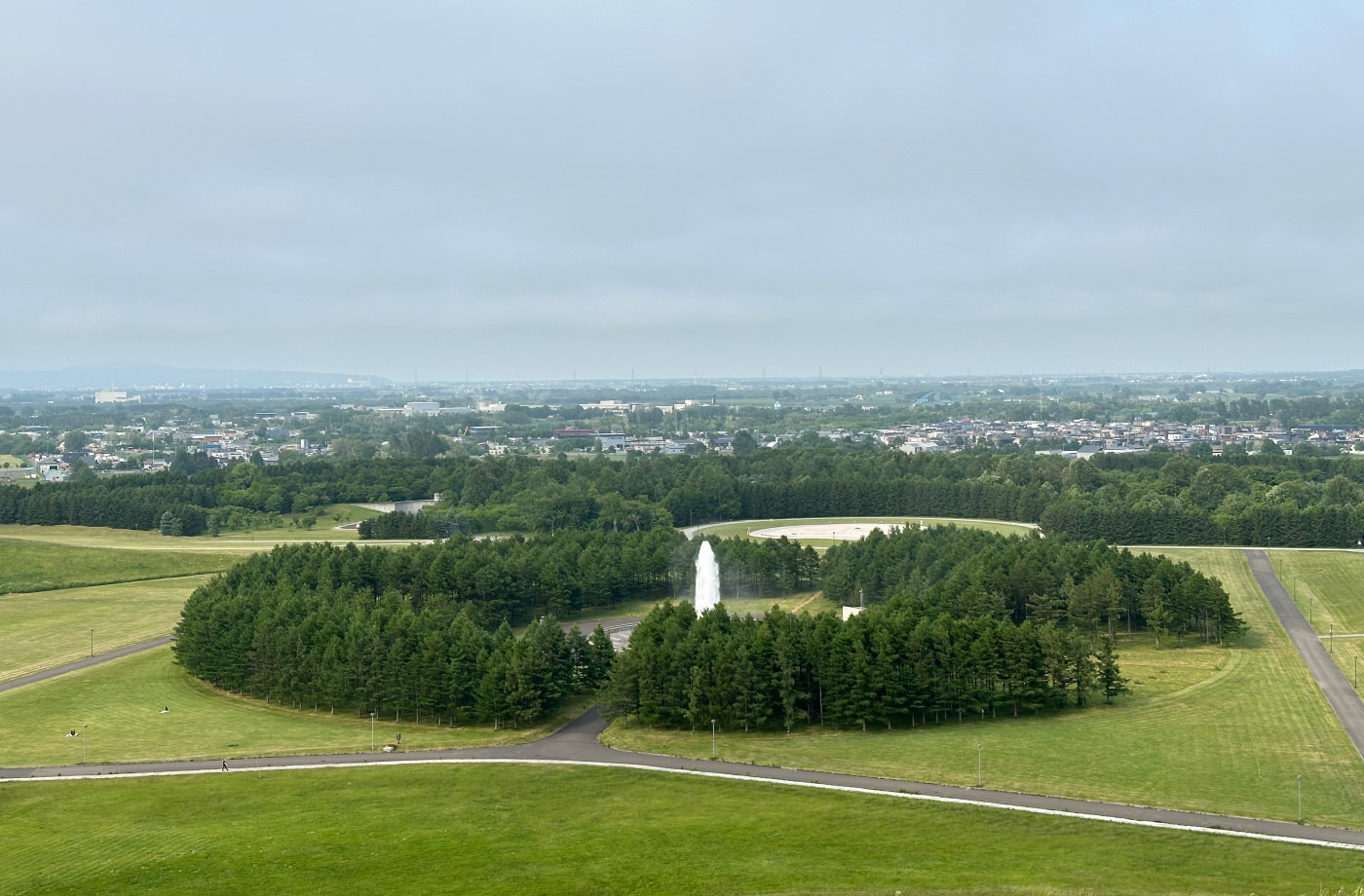 Milsvidd utsikt över en park där en cirkel med skog omger en fontän där vatten sprutar över 20 meter upp i luften. I fjärran bebyggelse och mer natur.