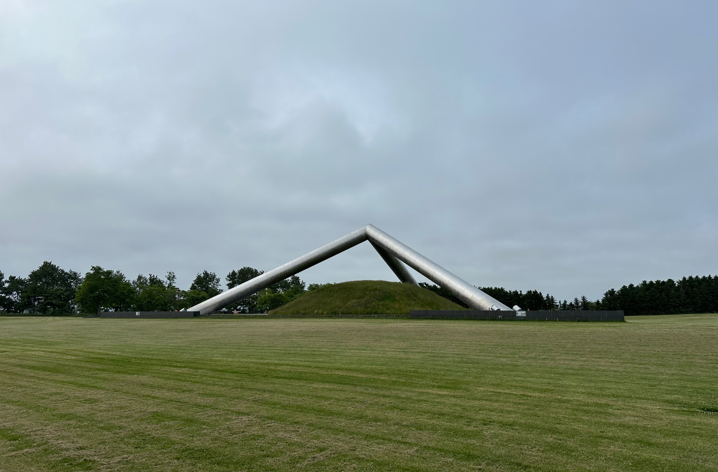 En många meter hög tetraeder byggd av tre cylinderformade ljusa metallrör. Den är omgiven av välklippt gräs.