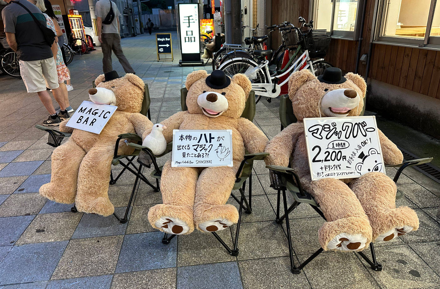 Tre stora nallar med små svarta hattar sitter i tre campingstolar med varsin skylt med japansk skrift. En lyder "Magic Bar".