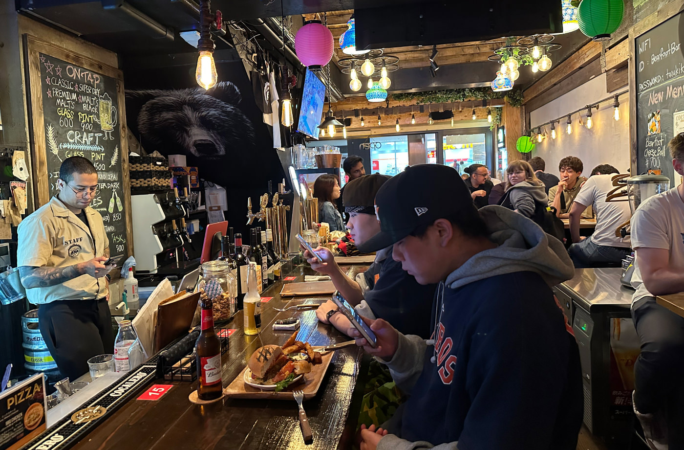 Välbesökt pub. Till vänster en bartender bakom bardisken, i mitten två gäster som tittar på sina telefoner och till höger fler göster som sitter vid bord.