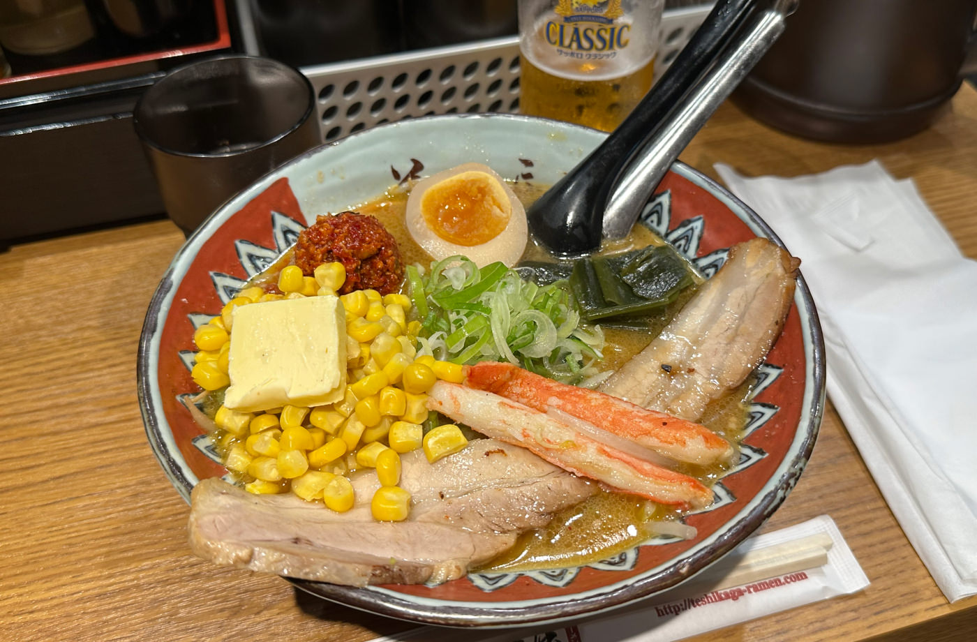 Stor skål med ramen med gräslök, krabba, majs, en smörklick, fläskkött och en ägghalva.