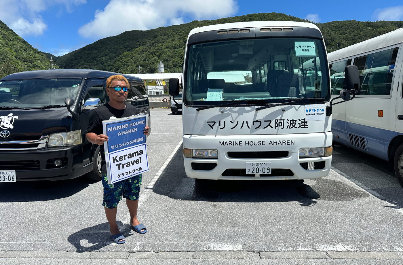 Tre bussar på en parkeringsplats. Bredvid den i mitten står en japansk man med gult hör, solglasögon och en skylt med texten "Kerama Travel".