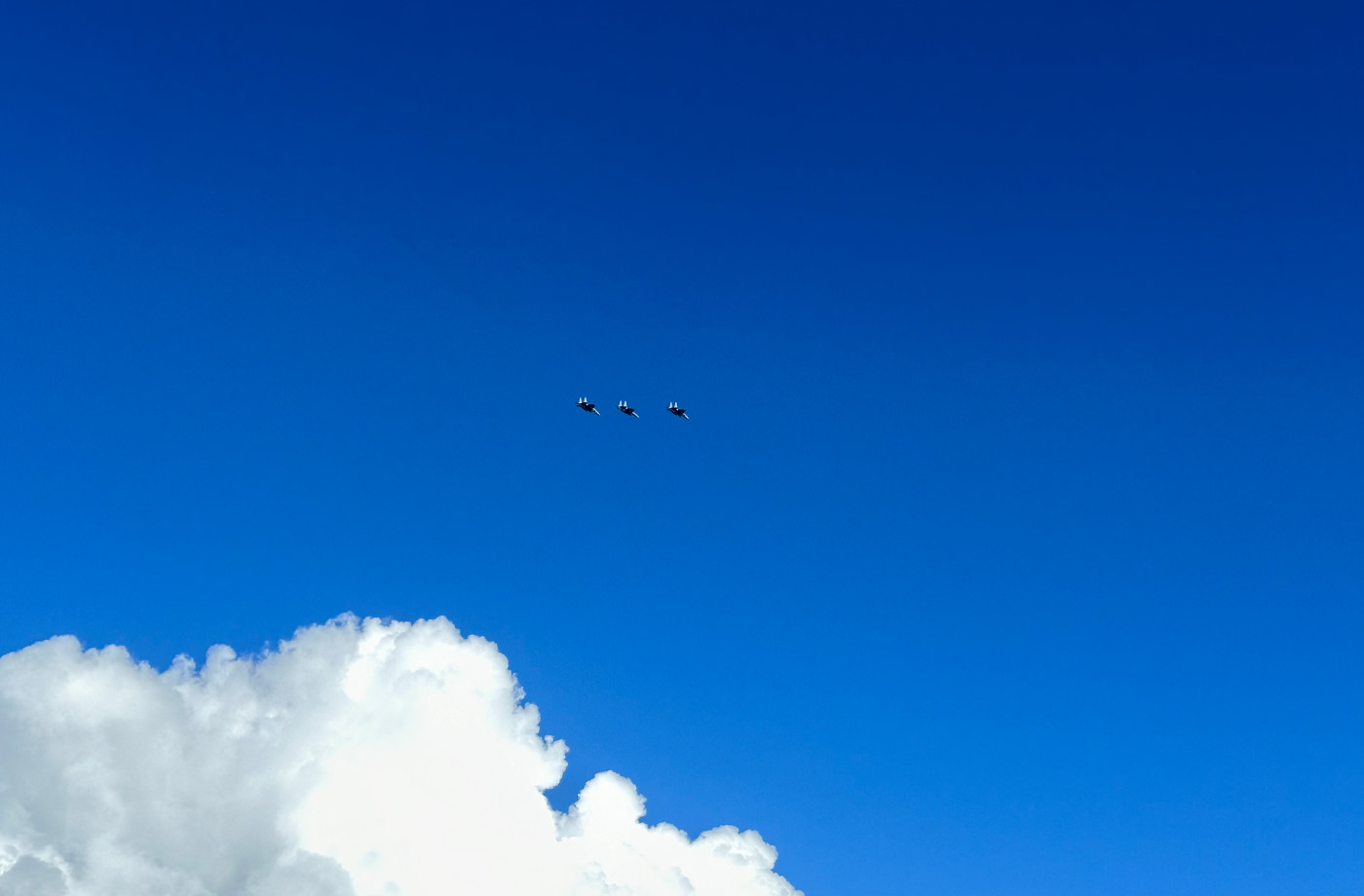 Tre stridsflygplan flyger i formation på en blå himmel. Ett stort vitt moln syns delvis i botten.