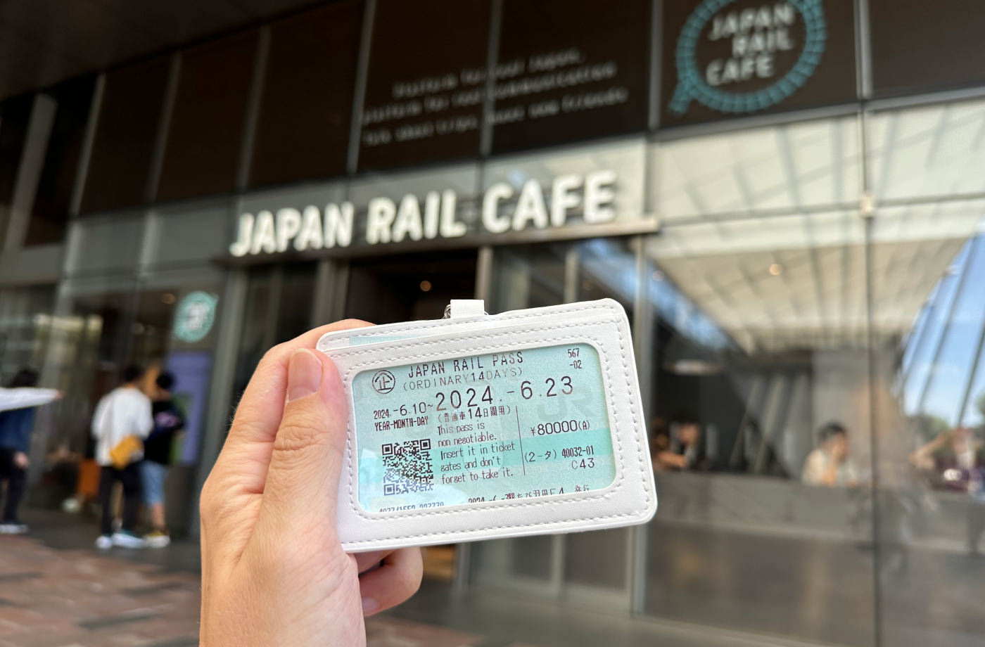 Hand håller i en ljusblå tågbiljett framför en byggnad med texten "Japan Rail Cafe".