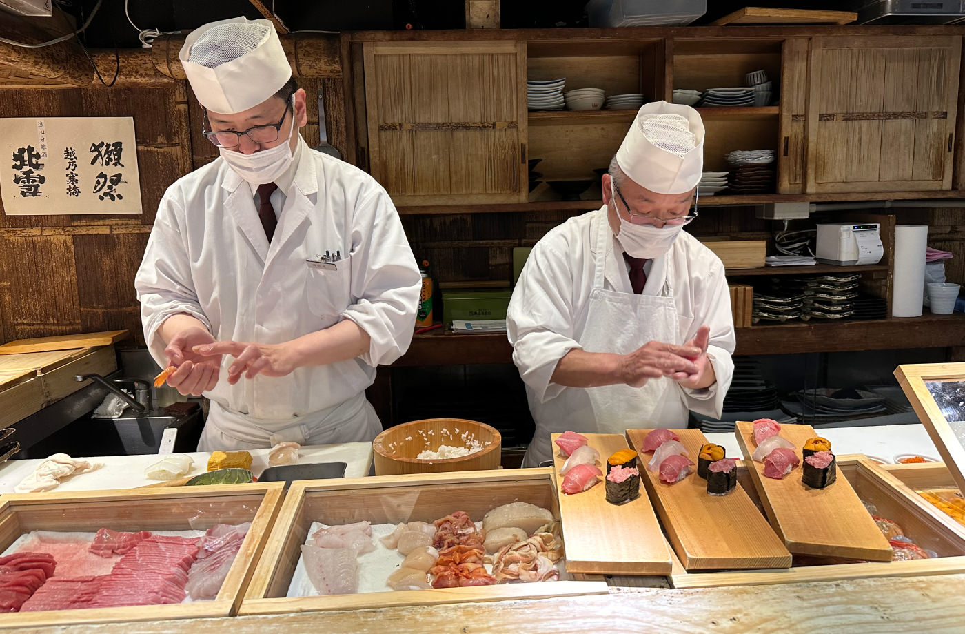 Två vitklädda sushikockar med munskydd står bakom en bardisk och gör klart tre tallrikar med sushi.