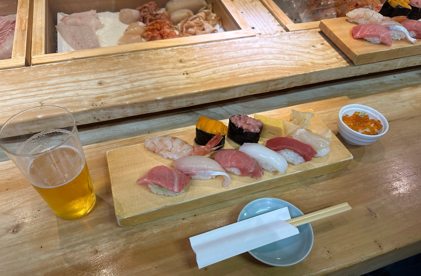 Nio bitar sushi på av rektangulär trätallrik på en bardisk. Även ett glas öl och en kopp med ris och laxrom.