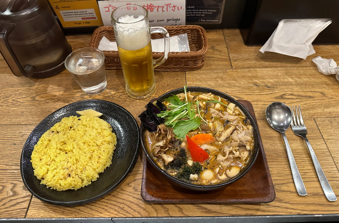 En tallrik med ris och en stor skål med currysoppa med grönsaker på en träbord. även ett glas vatten, ett stop öl och bestick.