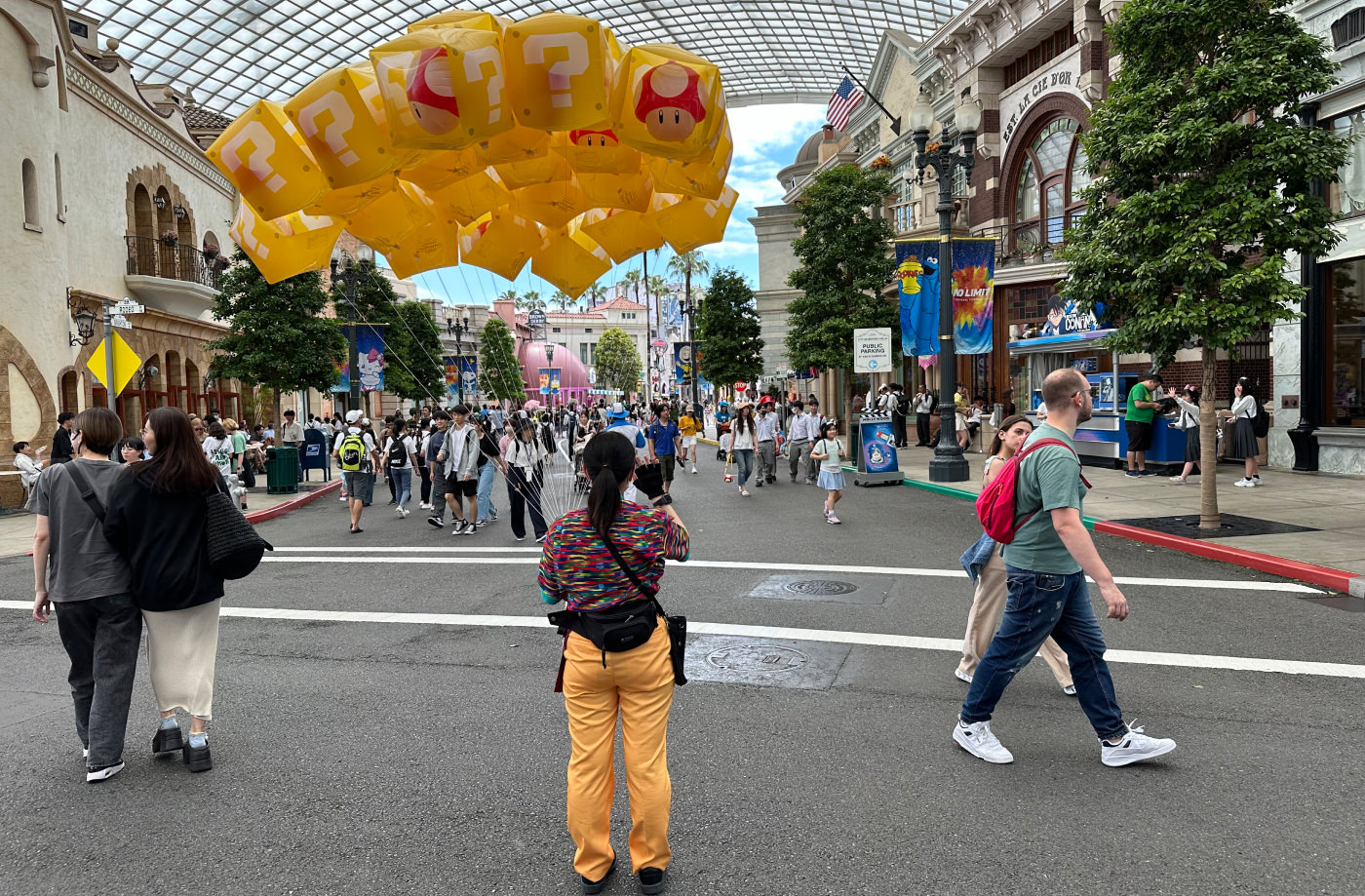 Kvinna i färgglada kläder stor på en gata med mycket folk på och håller i en drös med heliumballonger i trådar. Ballongerna är gula med vita frågetecken på.