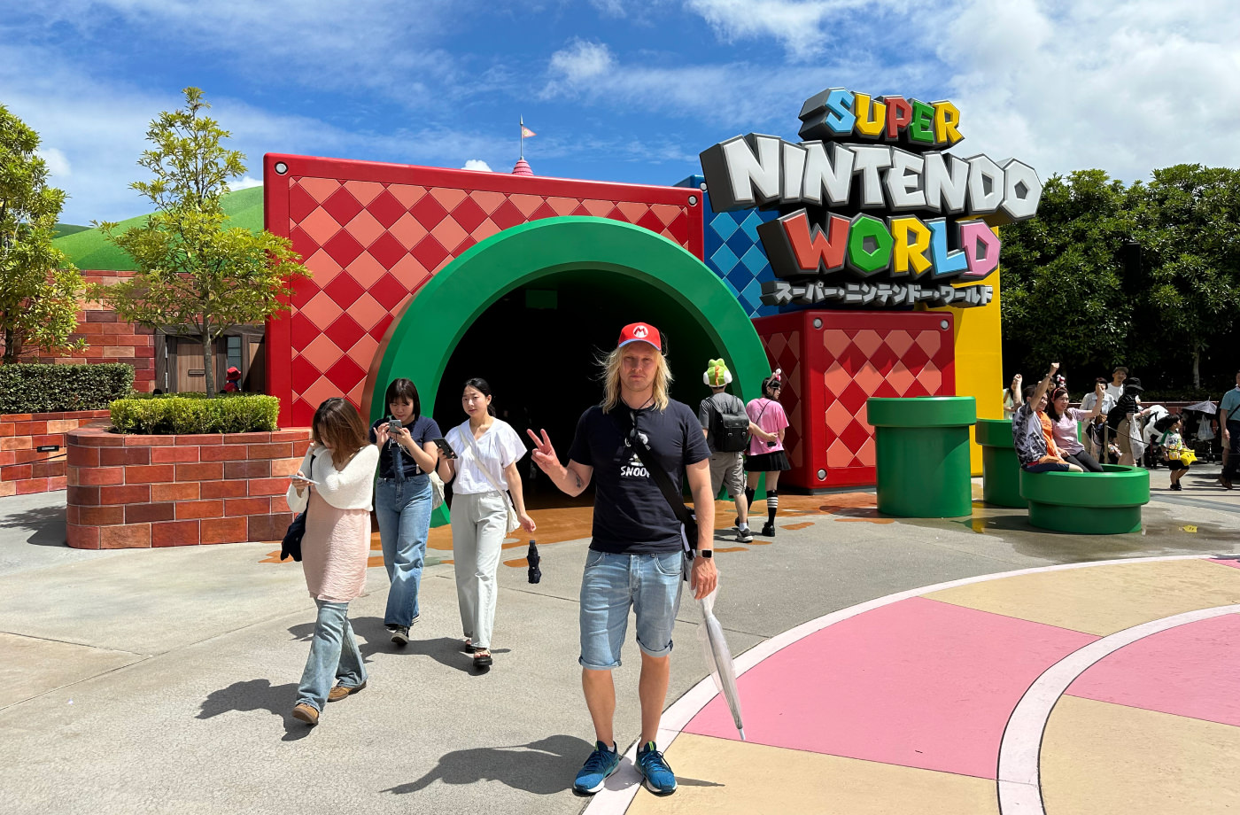 Jag står iförd shorts, t-shirt och keps framför en färgglad ingång i form av ett grönt rör från Super Mario-spelen. En skylt lyder "Super Nintendo World".