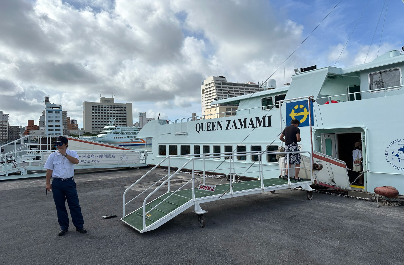 Vit snabbförja ankrad vid en pir. En ur personalen står vid en landgång och välkomnar folk. Text på färjan lyder "Queen Zamami".