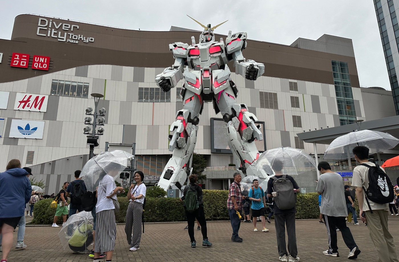 En enorm vit staty av en robot står i en park framför ett köpcenter. Vissa delar av robotens kropp lyser rött. Framför står personer med och utan paraplyn.