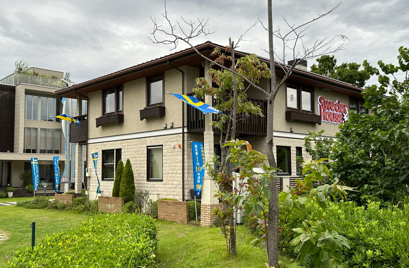 Ett ljust hus med brunt tak och bruna fönsterlister står i en lummig trädgård. En skylt på huset lyder "Sweden House". Från huset hänger flera svenska flaggor.