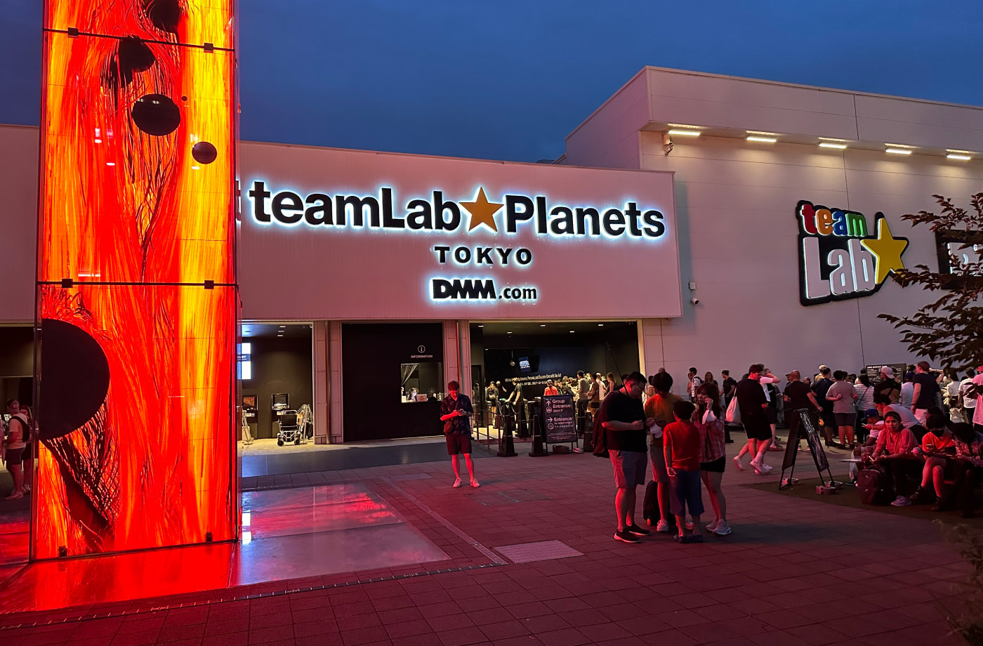 Stor vit byggnad med en upplyst skylt som lyder "teamLab Planets Tokyo". Till vänster står en stor glaspelare med videoskärmar som visar röd eld.