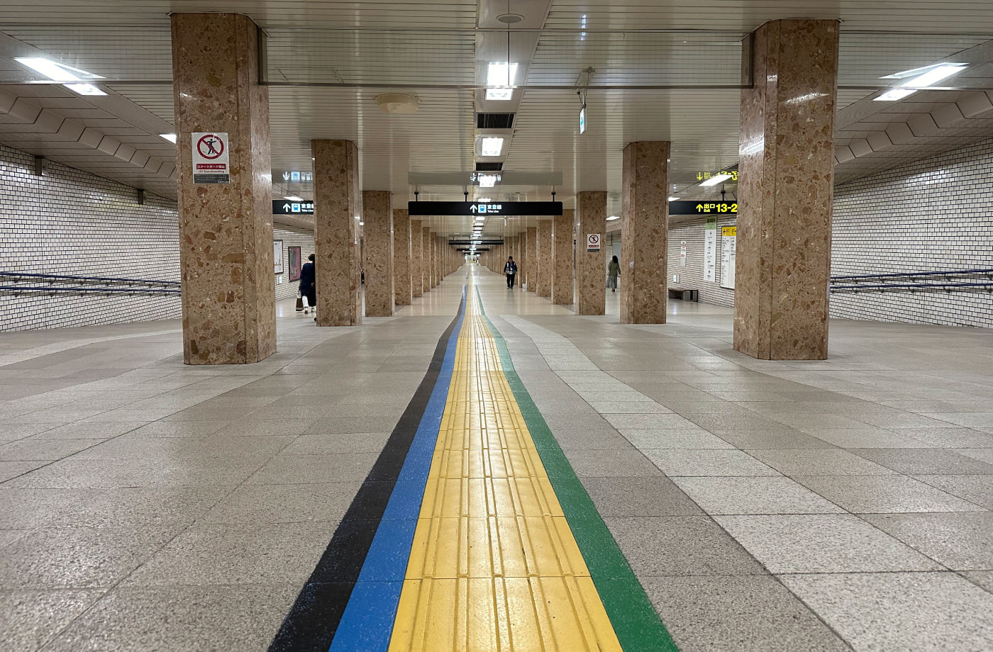 Underjordisk gång på en tågstation. På marken mitt emellan flera ljusbruna stenpelare är fyra linjer målade. En svart, blå, gul och grön.