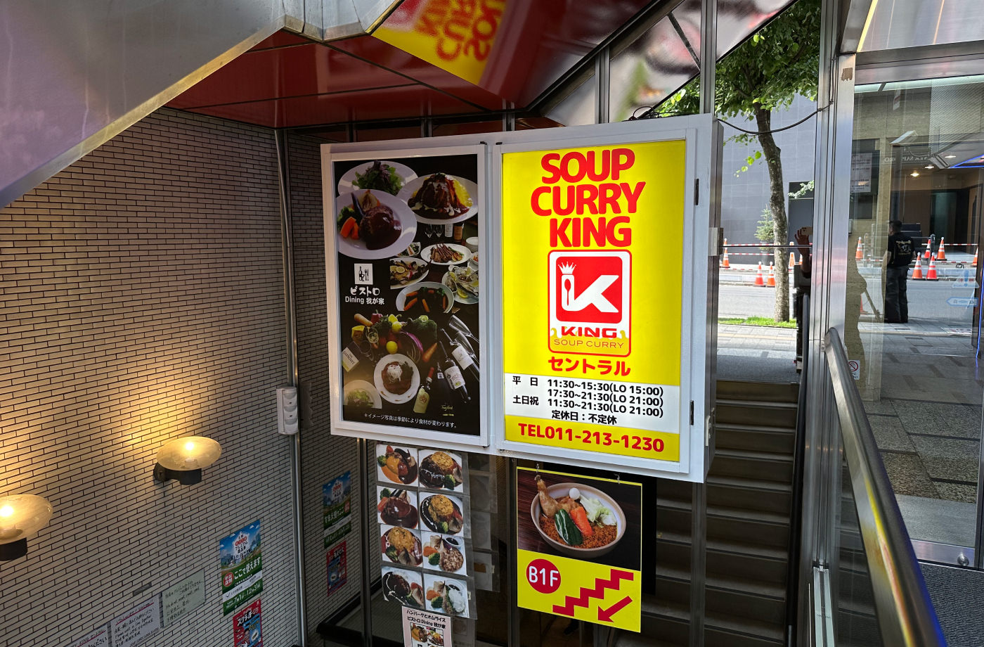 Två skyltar i en trappnedgång till en källare. Den ena är gul och röd med text som lyder "Soup Curry King". Den andra består av flera bilder på maträtter.